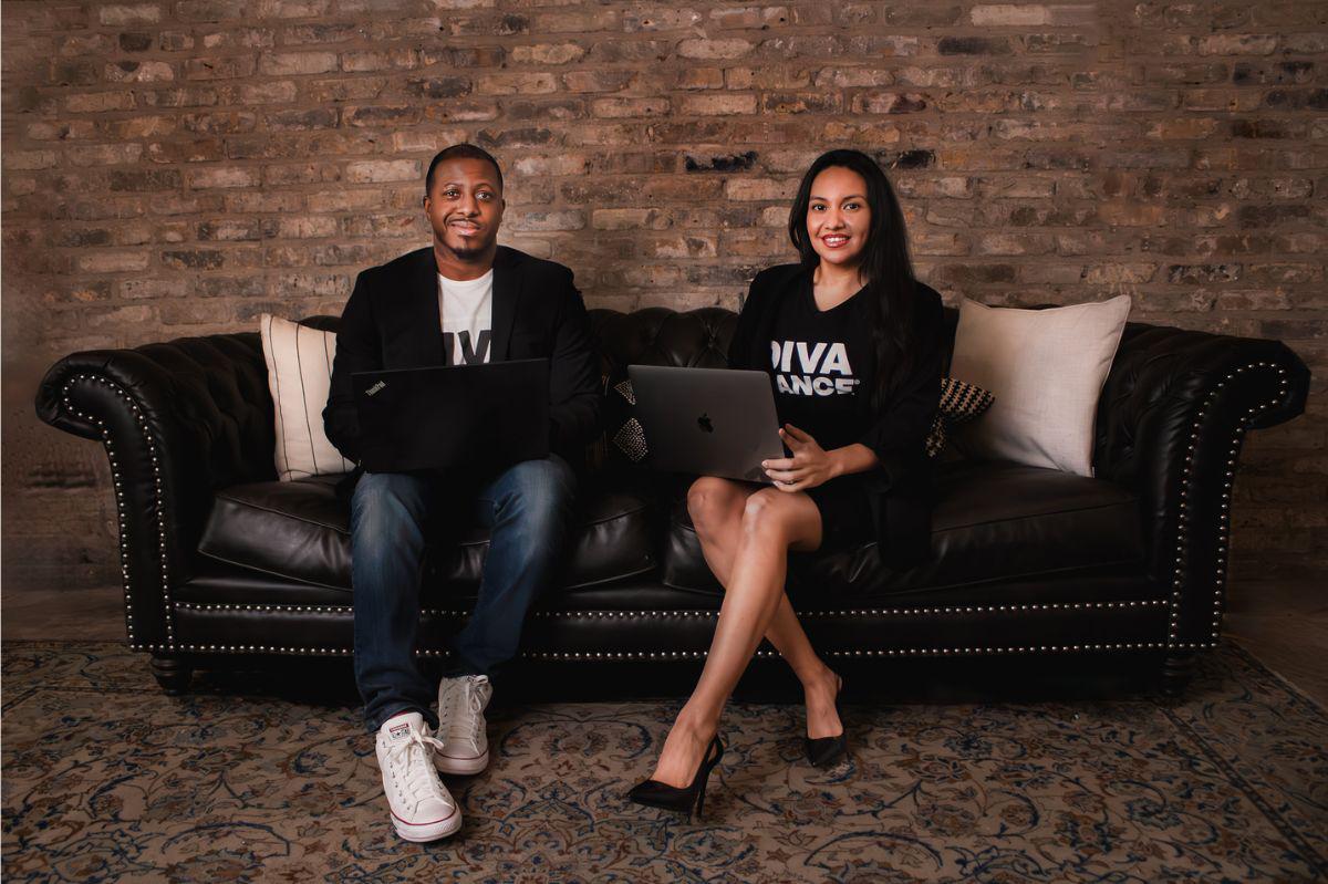 Two DivaDance leaders sit on a black sofa with laptops, smiling against a brick wall.
